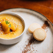 A beautifully plated crème brûlée with orange slices and shortbread biscuits, on a wooden table at the hotel restaurant.