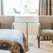 Cozy hotel room with plush chairs, glass table, patterned curtains, and warm lighting near a scenic window.