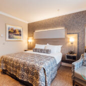 Elegant hotel room with king bed, plush armchair, modern decor, and soft lighting, offering a relaxing stay.